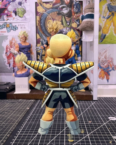 A Dragon Ball figure shown from the back, wearing a black and gold armor with light blue-gray boots and gloves, standing on a gridded cutting mat with anime posters in the background.