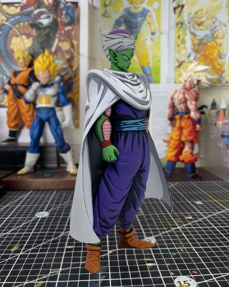 A detailed anime figurine of Piccolo Sempai from Dragon Ball Z, standing on a black gridded cutting mat with other collectible figures blurred in the background.