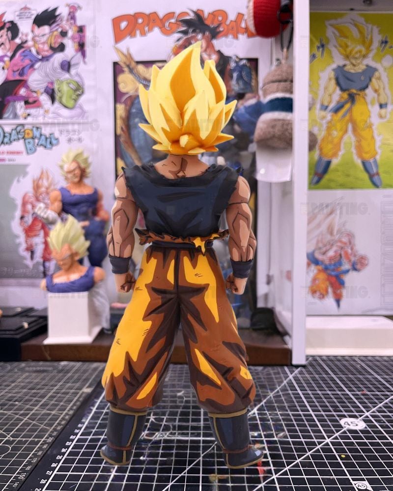 A Dragon Ball figure of Goku in a Super Saiyan form, viewed from behind, standing on a black gridded cutting mat in a collector's room with Dragon Ball posters in the background.