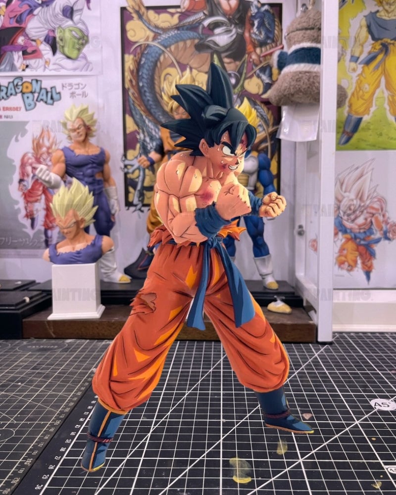 A Dragon Ball figure of Goku in a fighting stance, posed on a black gridded cutting mat with various collectibles and posters in the background.