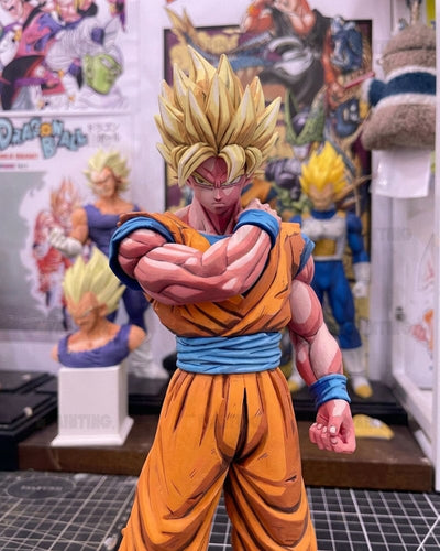 A detailed collectible figurine of Goku in a Super Saiyan form, wearing his iconic orange and blue Dragon Ball Z outfit, posed with arms crossed in a workshop or collector's room with Dragon Ball posters in the background.
