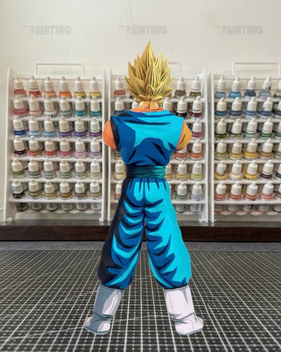 A hand-painted anime figure of Vegetta from Dragon Ball Z, shown from the back wearing his blue and orange gi with spiky blond hair, standing on a cutting mat in front of shelves of paint bottles.
