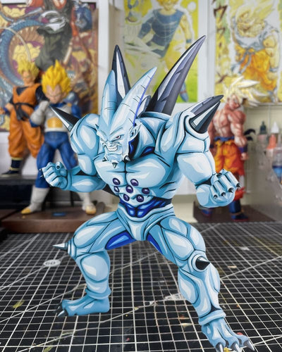 A light blue and white anime action figure of a muscular horned monster, posed on a gridded cutting mat with blurred Dragon Ball Z character figures in the background.