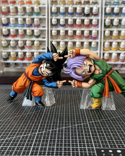 Two Dragon Ball Z anime figurines, Goku and Trunks, posed in a fighting stance on clear plastic stands over a gridded cutting mat, with shelves of miniature paint bottles in the background.
