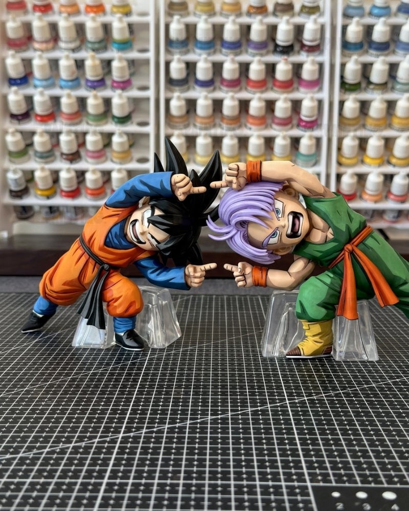 Two Dragon Ball Z anime figurines, Goku and Trunks, posed in a fighting stance on clear plastic stands over a gridded cutting mat, with shelves of miniature paint bottles in the background.