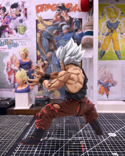 A detailed collectible action figure of Goku in a battle stance, featuring spiky silver hair, a torn black top, and torn brown pants, displayed on a gridded cutting mat with Dragon Ball posters in the background.