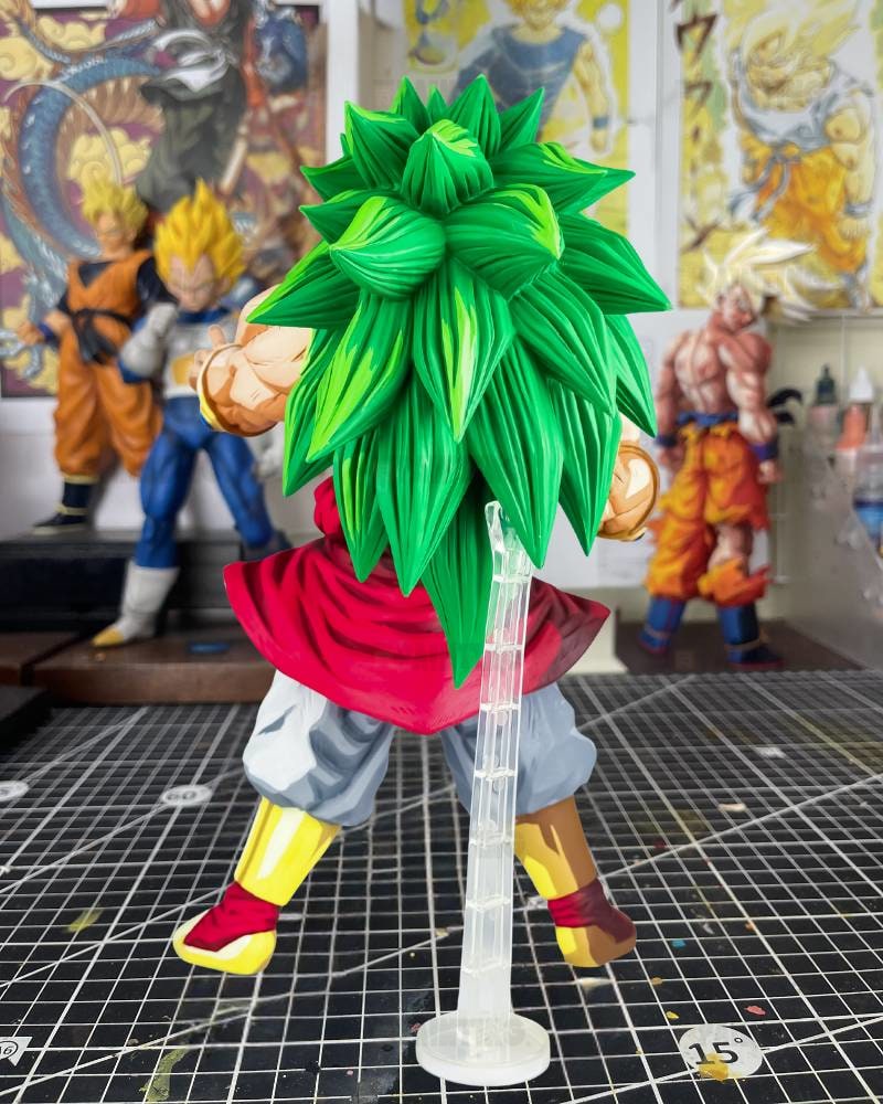 A Dragon Ball Z anime figure with a large green spiky hair style, wearing a red top, gray pants, and yellow boots, standing on a clear display stand on a gridded cutting mat with other collectible figures in the background.