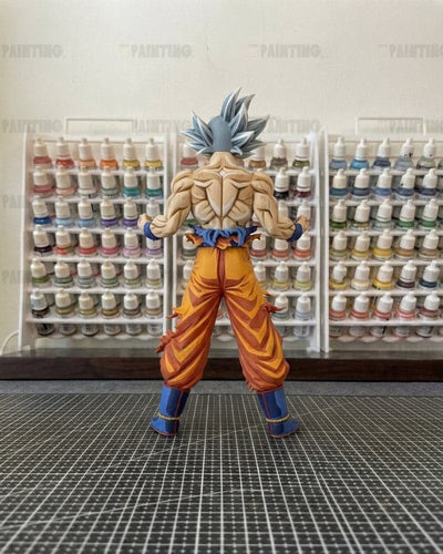 A painted Dragon Ball figure of Goku from the back, showcasing his spiky blue hair, muscular back, orange gi pants, and blue boots. The figure is displayed on a black grid cutting mat with shelves of miniature paint bottles in the background.