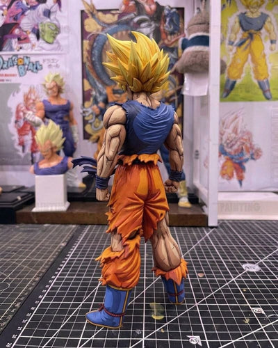 A detailed anime action figure of Goku in a Super Saiyan form, shown from the back on a black gridded cutting mat with Dragon Ball themed artwork in the background.