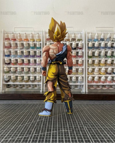 A detailed anime action figure of Goku in a Super Saiyan pose, standing on a black gridded cutting mat with shelves of miniature paint bottles in the background.