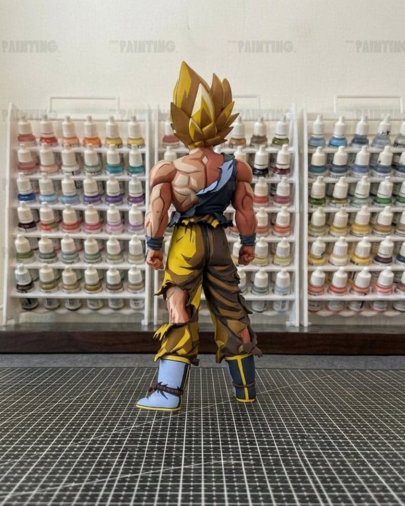 A detailed anime action figure of Goku in a Super Saiyan pose, standing on a black gridded cutting mat with shelves of miniature paint bottles in the background.