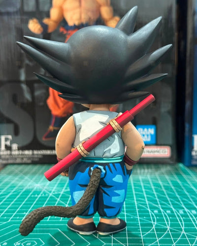 A Dragon Ball figure shown from the back, featuring spiky black hair, a white sleeveless top, blue patterned pants, a red sai weapon, and a long dark tail, standing on a green cutting mat with a blurred collectible box in the background.