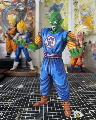 A detailed collectible figurine of the character Piccolo from Dragon Ball Z, standing on a black grid cutting mat in a hobby room with other Dragon Ball collectibles in the background.