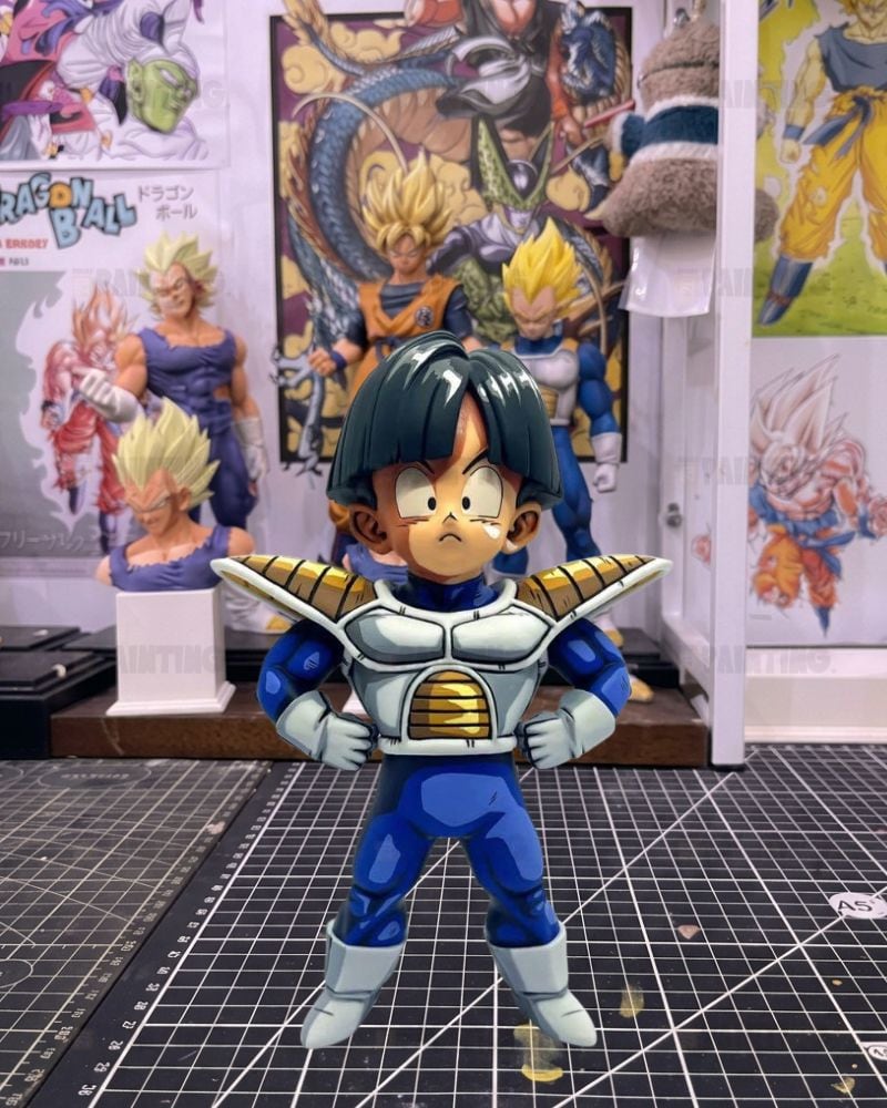 A Dragon Ball figure of a young Goku in a blue and white battle suit with gold accents, standing on a gridded cutting mat in front of posters and other collectibles.