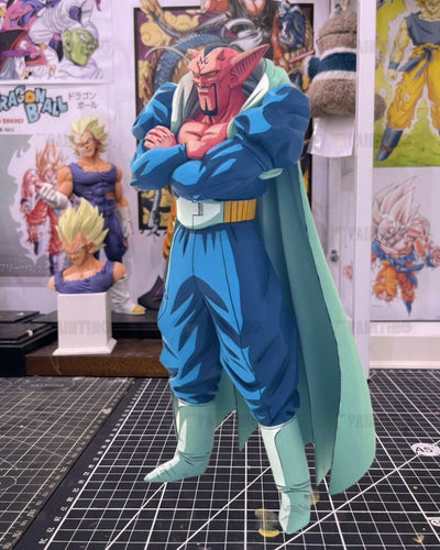 A Dragon Ball figure of a pink-skinned warrior with horns, wearing a blue outfit and light green cape, standing on a black gridded cutting mat with anime posters in the background.
