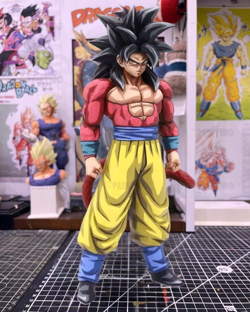 A detailed anime figurine of Goku in his Super Saiyan form, standing on a black gridded cutting mat with Dragon Ball posters and a display stand in the background.