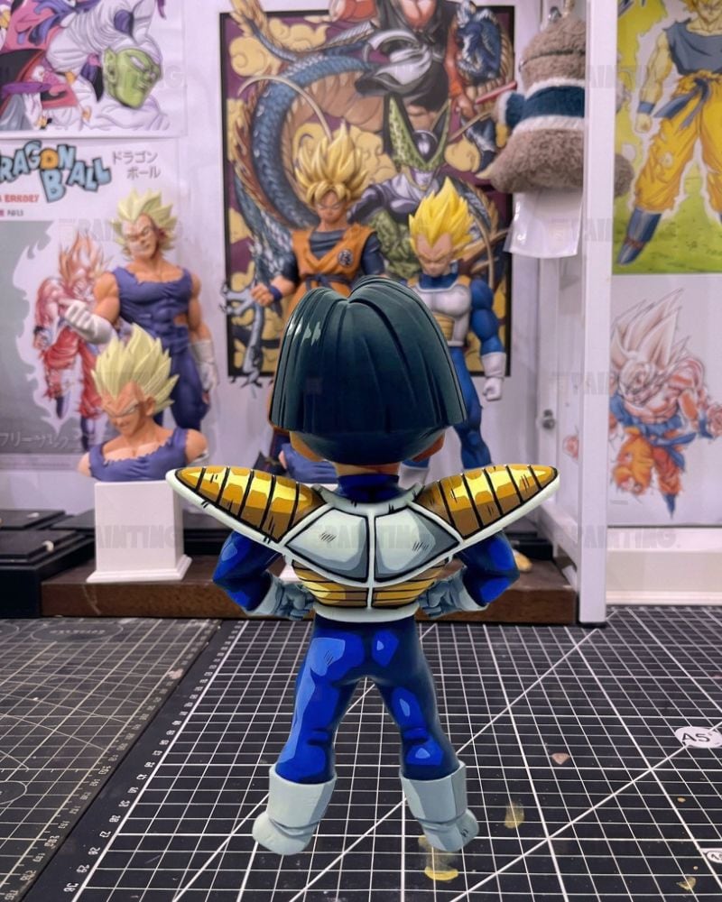 A Dragon Ball figure shown from the back, wearing a blue and white outfit with gold shoulder armor, standing on a gridded cutting mat in front of Dragon Ball posters.