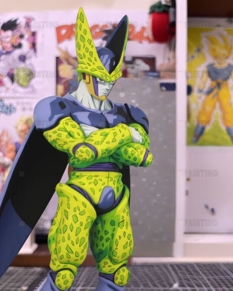 A Dragon Ball figure of Cell with arms crossed, standing on a gray grid surface in front of a wall with anime posters.