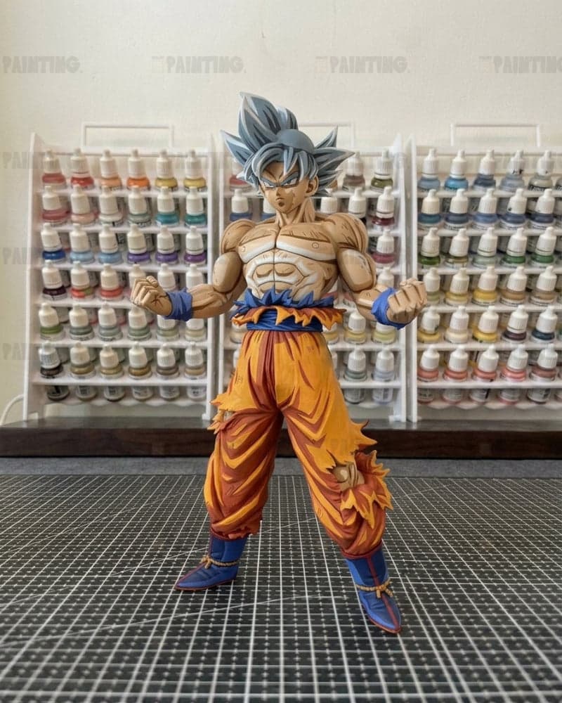 A detailed anime figurine of Goku in a fighting pose, standing on a black gridded mat with shelves of paint bottles in the background.