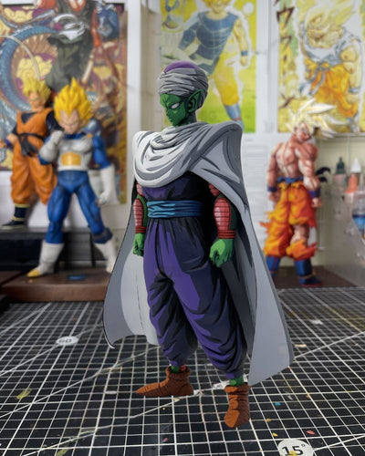 A Dragon Ball Z collectible figure of Piccolo standing on a gridded cutting mat, with other anime figures and posters blurred in the background.