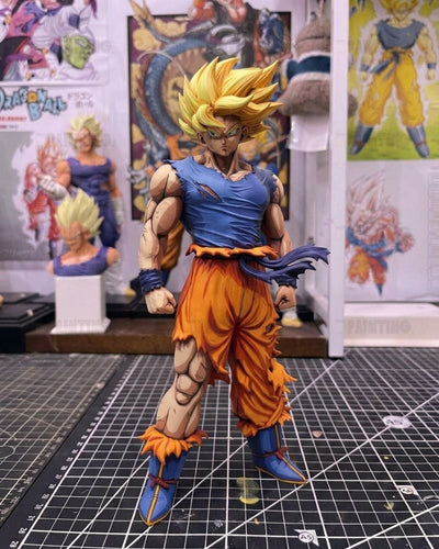 A detailed anime figurine of Goku in a Super Saiyan transformation, standing on a black gridded cutting mat with Dragon Ball collectibles in the background.