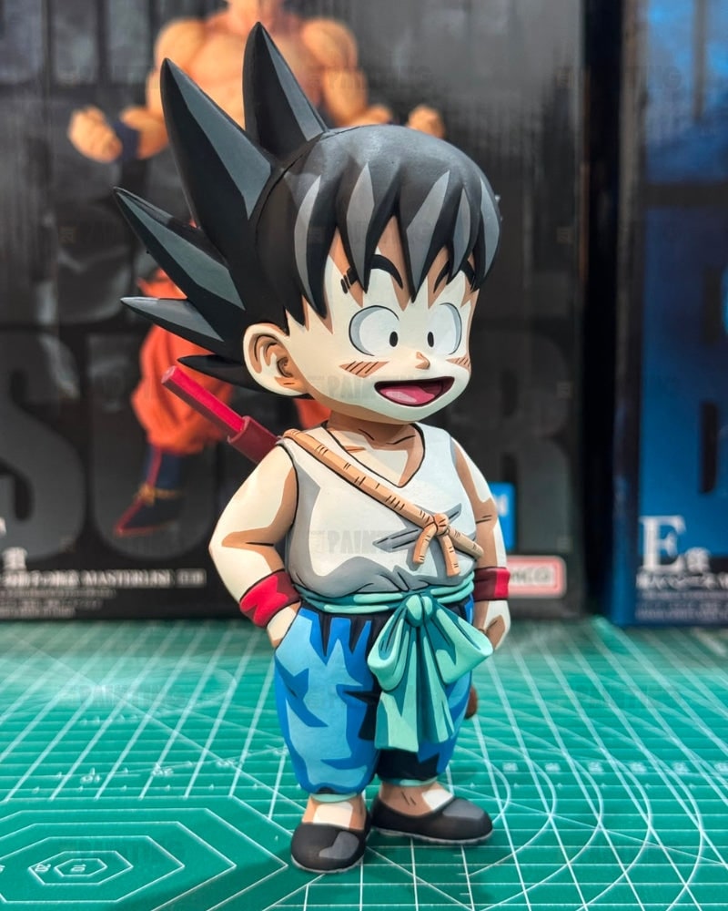 A chibi-style anime figurine of Goku standing on a green cutting mat, with Dragon Ball Z packaging boxes blurred in the background.