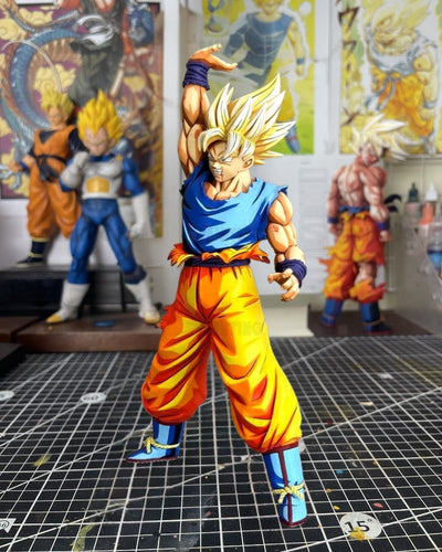 A detailed anime figurine of Goku in his Super Saiyan form, posed dynamically on a gridded cutting mat with other Dragon Ball collectibles in the background.