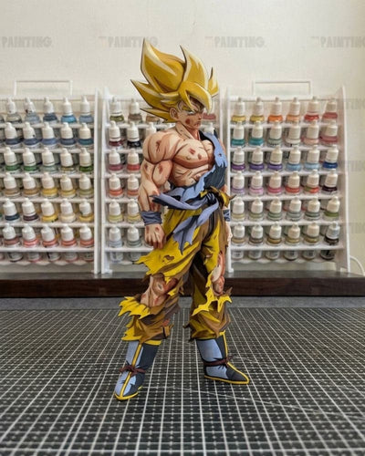 A detailed anime figurine of a Super Saiyan character, standing on a black gridded cutting mat with shelves of small paint bottles in the background.