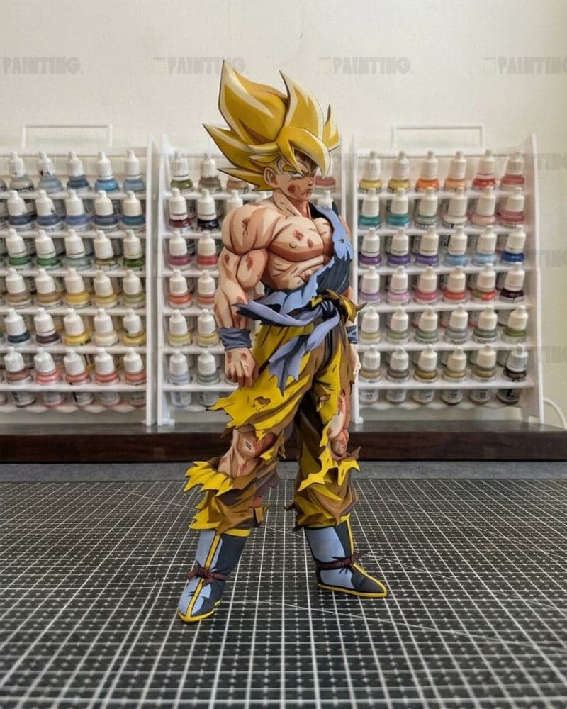A detailed anime figurine of a Super Saiyan character, standing on a black gridded cutting mat with shelves of small paint bottles in the background.
