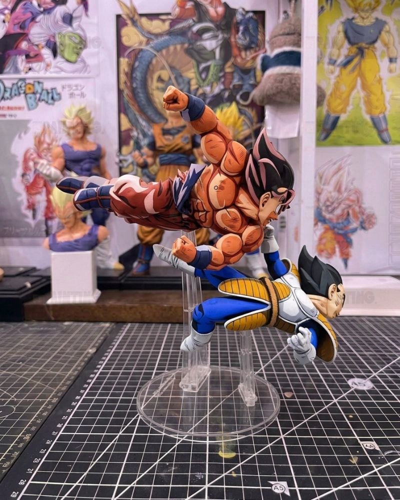 Dragon Ball figure of Goku performing a flying kick over Vegeta on a clear acrylic stand, set on a gridded cutting mat with Dragon Ball collectibles in the background.