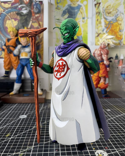 A detailed collectible action figure of Piccolo from Dragon Ball Z, standing on a black gridded cutting mat in a hobby room. The figure features his signature green skin, white robe with a red Chi symbol, purple collar, and holds a brown staff.