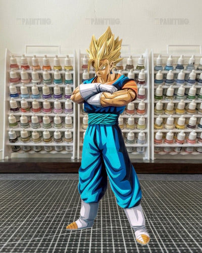 A die-cut sticker or decal of the anime character Vegetta from Dragon Ball Z, standing on a black gridded cutting mat with shelves of paint bottles in the background.