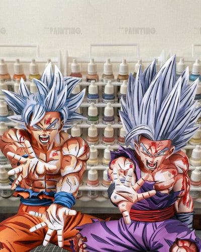 Two anime-style collectible figures resembling Super Saiyan Gohan from Dragon Ball Z, posed in front of a wall of paint bottles. The figure on the left wears orange pants and a blue sash, while the figure on the right wears a purple gi with a red sash.