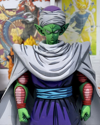 A collectible statue of the anime character Piccolo from Dragon Ball Z, featuring his signature green skin, purple and blue outfit, and white head wrap, set against a blurred background of colorful Dragon Ball posters.