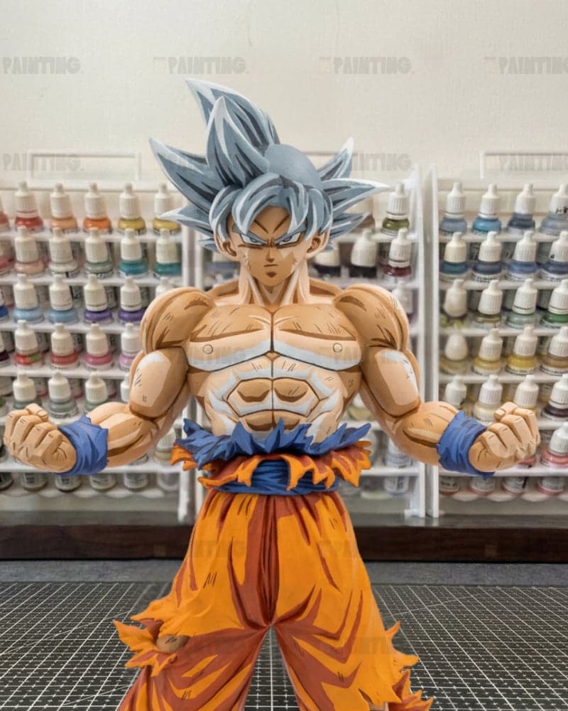 A painted anime figurine of Goku in his Super Saiyan form, standing in a workshop with shelves of paint bottles in the background.