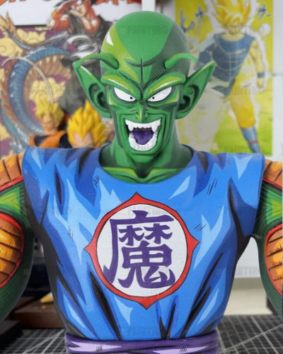 A collectible bust statue of Piccolo from Dragon Ball Z, featuring his signature green skin and blue gi with the Dragon Ball symbol on the chest, set against a blurred background of Dragon Ball posters.