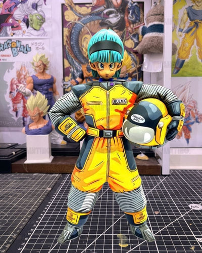 A Dragon Ball figure of Bulma in her signature yellow and gray jumpsuit, holding a yellow and gray helmet, standing on a cutting mat with Dragon Ball collectibles in the background.