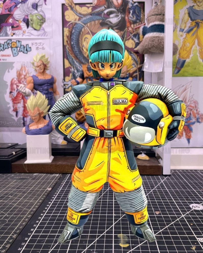 A Dragon Ball figure of Bulma in her signature yellow and gray jumpsuit, holding a yellow and gray helmet, standing on a cutting mat with Dragon Ball collectibles in the background.