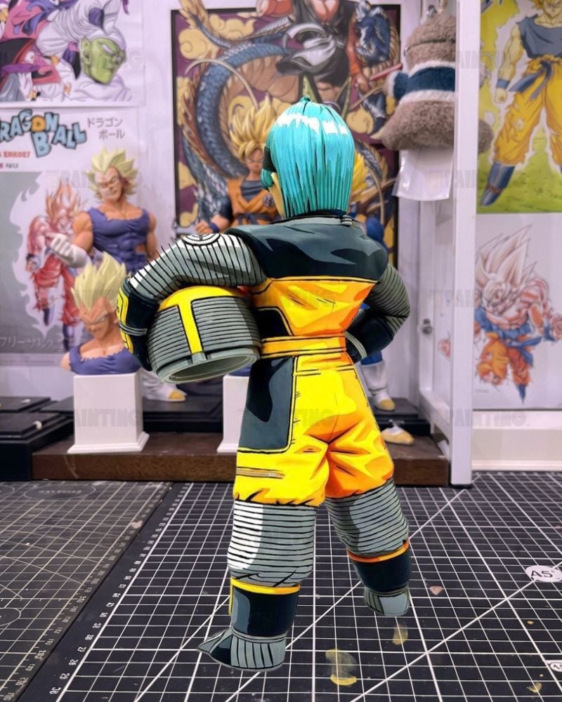 A Dragon Ball figure shown from the back, wearing a yellow and dark gray outfit with bright blue hair, standing on a gridded cutting mat in front of Dragon Ball posters.