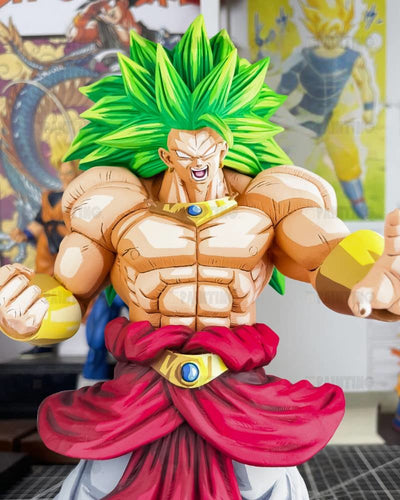 A detailed collectible figurine of Broly from Dragon Ball Z, featuring his classic green hair, muscular physique, red sash, and gold chest piece, set against a blurred background with Dragon Ball posters.