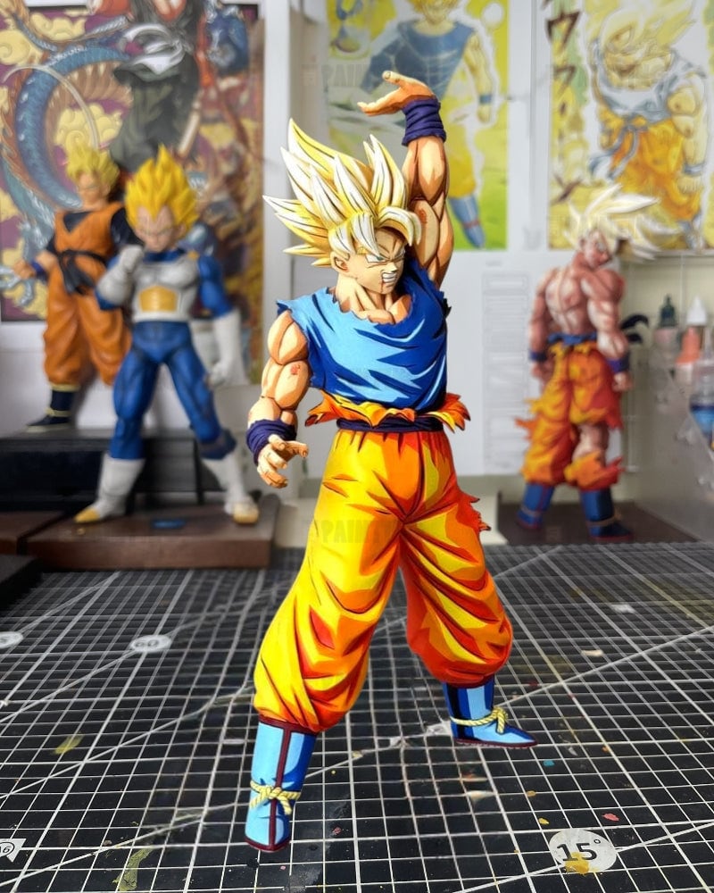 A detailed anime figurine of Goku in a Super Saiyan pose, wearing his classic blue and orange gi, standing on a black gridded cutting mat with other Dragon Ball collectibles in the blurred background.