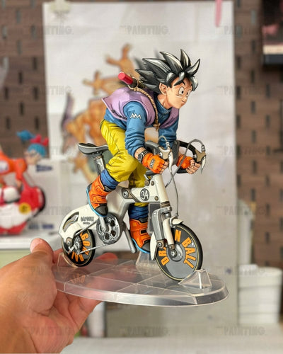 A hand holds a detailed anime figurine of Goku riding a futuristic bicycle on a clear acrylic stand. The figurine features spiky black hair, a blue and purple jacket, yellow pants, and orange boots, set against a blurred indoor background.
