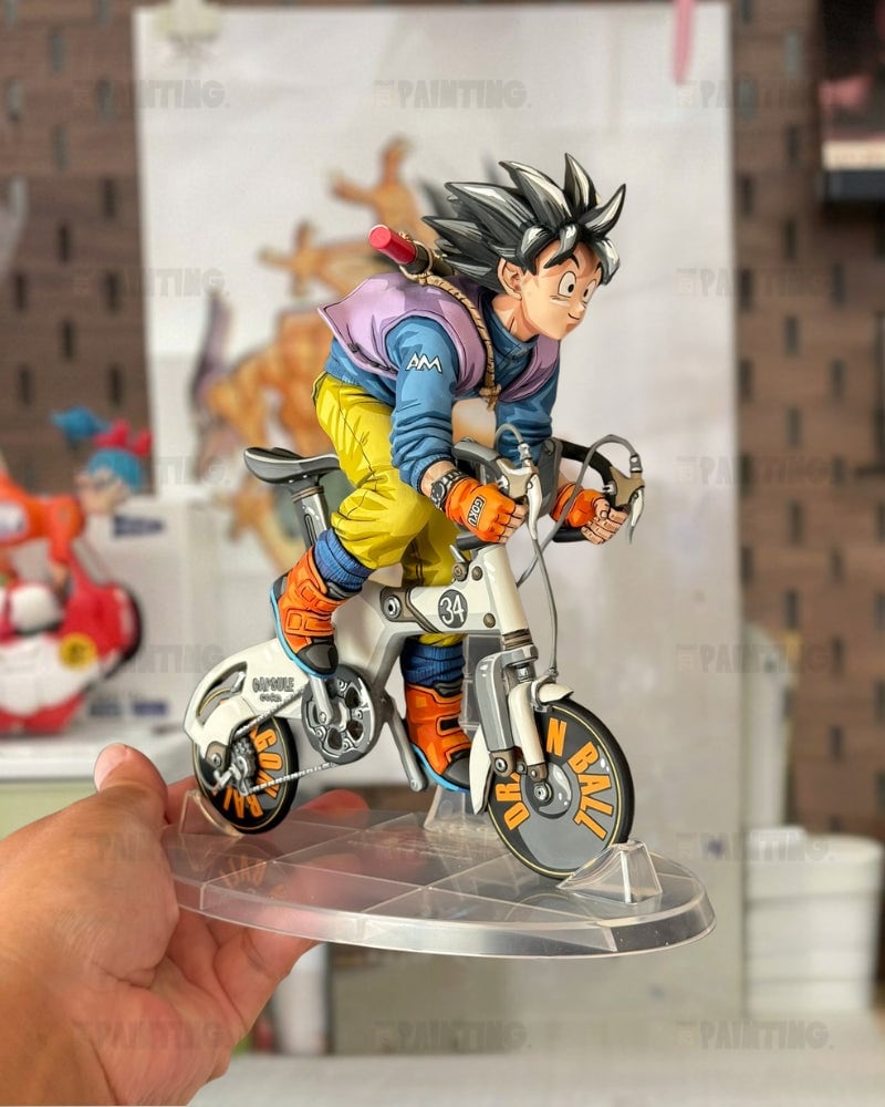 A hand holds a detailed anime figurine of Goku riding a futuristic bicycle on a clear acrylic stand. The figurine features spiky black hair, a blue and purple jacket, yellow pants, and orange boots, set against a blurred indoor background.