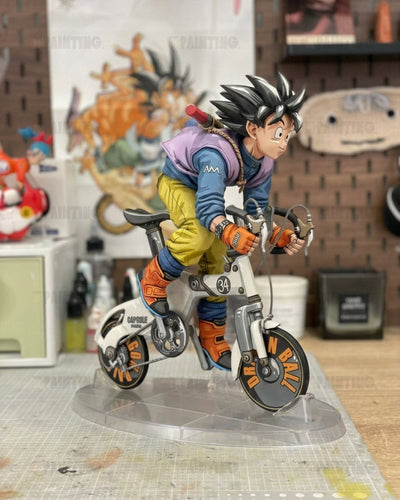 A detailed anime figurine of Goku riding a small white bicycle on a clear acrylic stand, set against a blurred workshop background.