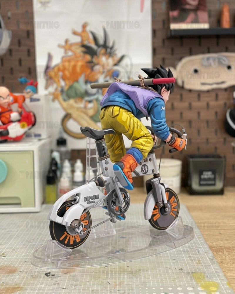 A detailed anime figurine of Goku riding a white bicycle, displayed on a clear acrylic stand in a collector's room setting.