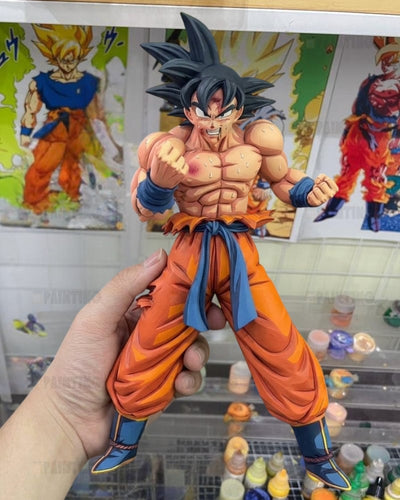 A hand holds a detailed anime figurine of Goku from Dragon Ball Z, wearing his signature orange gi and blue sash, with posters of Goku in the background.