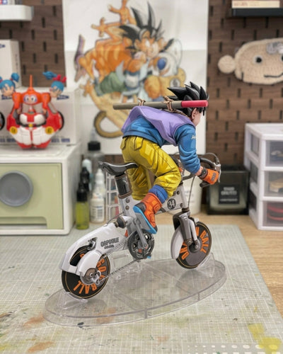 A detailed anime figurine of Goku riding a white electric bicycle, displayed on a clear acrylic stand in a collector's room setting.